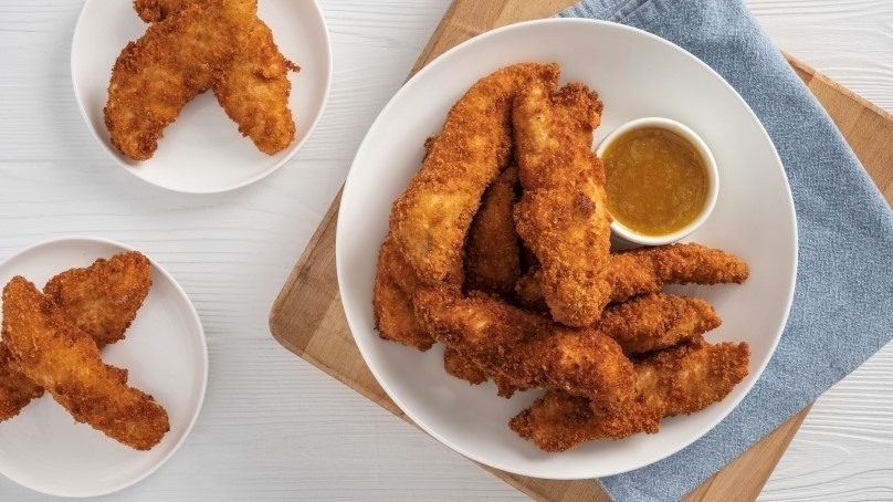 Crunchy Baked Parmesan Chicken Strips on white plate with dipping sauce, blue napkin, cutting board, white wood table