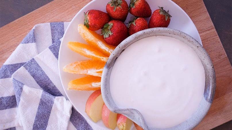 Cream Cheese Marshmallow Fruit Dip in bowl with sliced apples, orange slices, strawberries, kithcne towel, cuting board