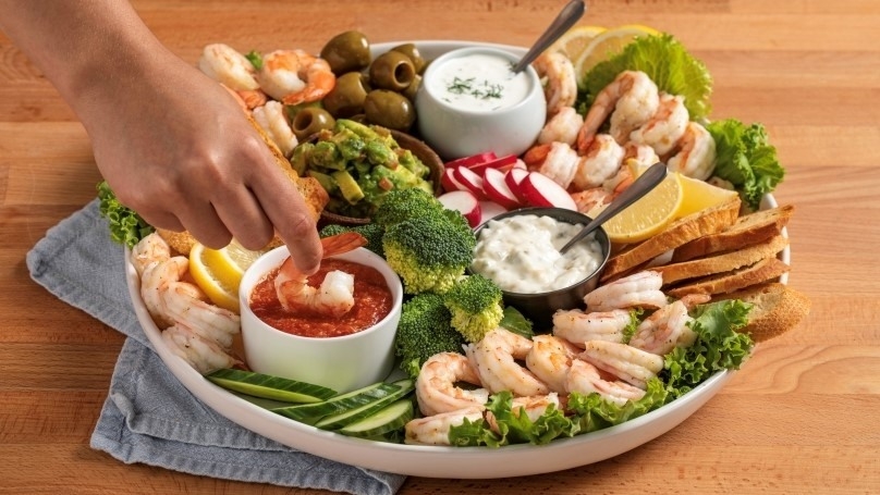 Hand dipping shrimp in cocktail sauce, snack board with shrimp, lemons, cucumbers, radishes, guacamole, fresh baguette slices, tzatziki dip, tartar sauce, etc.