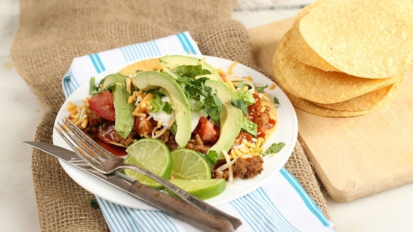 Beef Tostadas, salsa, sliced avocado, shredded cheese, cilantro, knife and fork, white plate, stiped kitchen towel, tortillas on cutting board, woven tablecloth