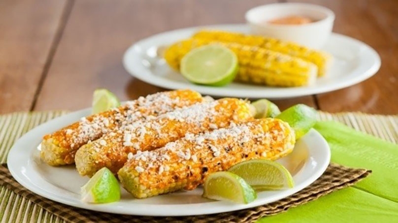 Mexican Grilled Corn on plate with queso fresco and lime wedges, woven placemat, wood table