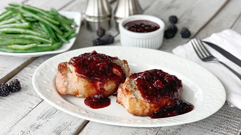 Grilled Pork chops with blackberry glaze on white plate, green beans, blackberries, salt and pepper mills, cruent of glaze, napkin, knife and firk, light gray wood table