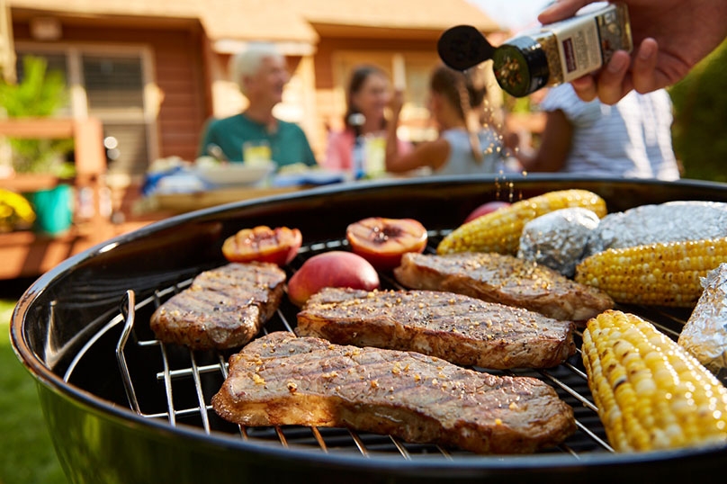 Steaks, corn, peaches and potatoes cooking on the grill, hand shaking seasoning on steaks, Family and picnic table in background