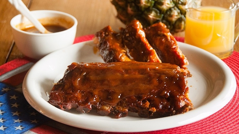 Apricot Pineapple Ribs on white plate, serving bowl of BBQ sauce, fresh pineapple, cup of pineapple juice, red placemat, wood table