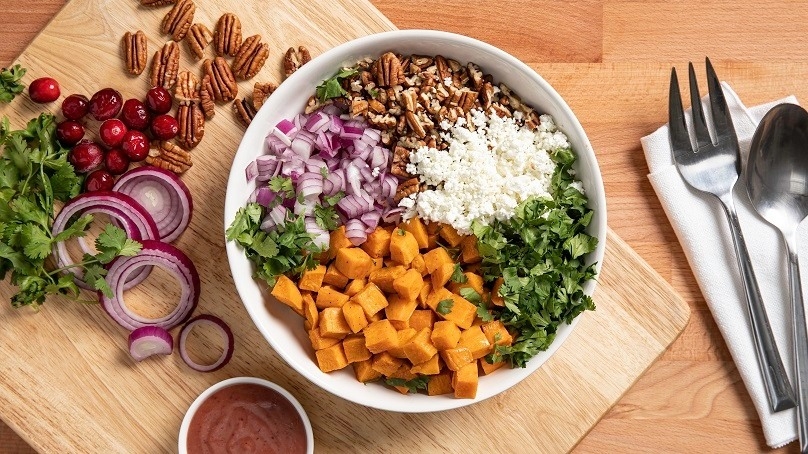 Roasted Sweet Potato Salad in bowl, cutting board, pecans, cranberries, sliced red onion, parsely, dressing in cruet, wood table