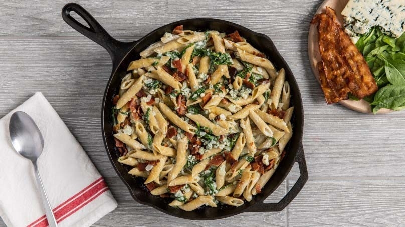 Asparagus and mushroom carbonara in iron pan with bacon, bleu cheese, and fresh spinach on plate, red and white napkin, spoon, gray wood table