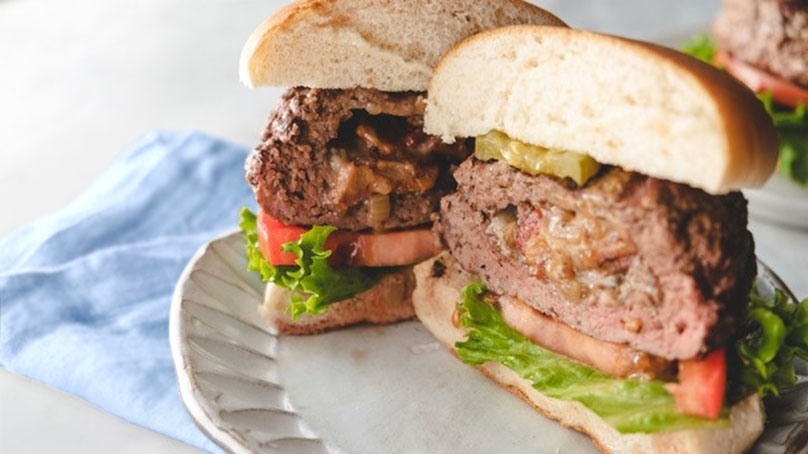 Closeup of halved grilled bacon stuffed burger, white plate, blue napkin, white counter