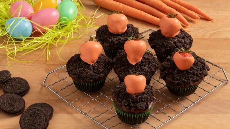 Easter Carrot Patch Chocolate Cupcakes, topped with candy dipped strawberries, chocolate sandwich cookies, easter eggs on artificial grass, carrots, wood table