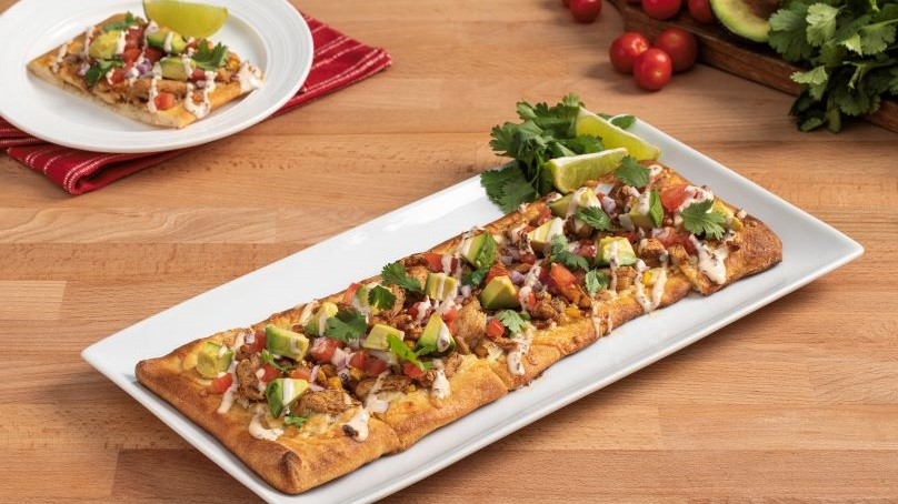 Plant-based chipotle chicken flatbread pizza with avocado, tomatoes, cheese and a chipotle mayo drizzle on square plate with lime and fresh cilantro garnish