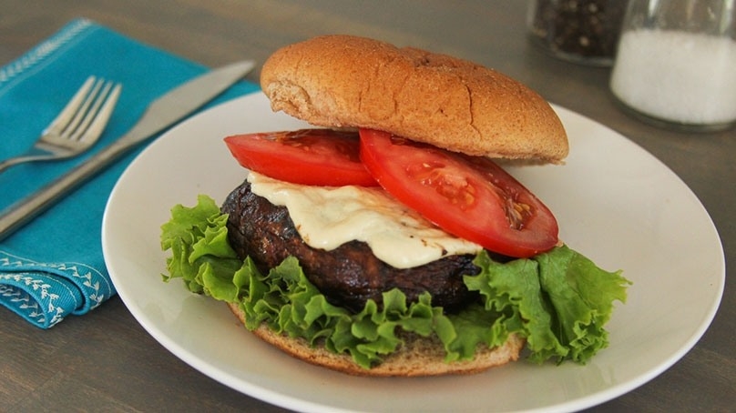 Portabella mushroom burger with lettuce, cheese, and tomato on white plate, blue dinner napkin with fork and knife, salt and pepper shakers, gray wood table