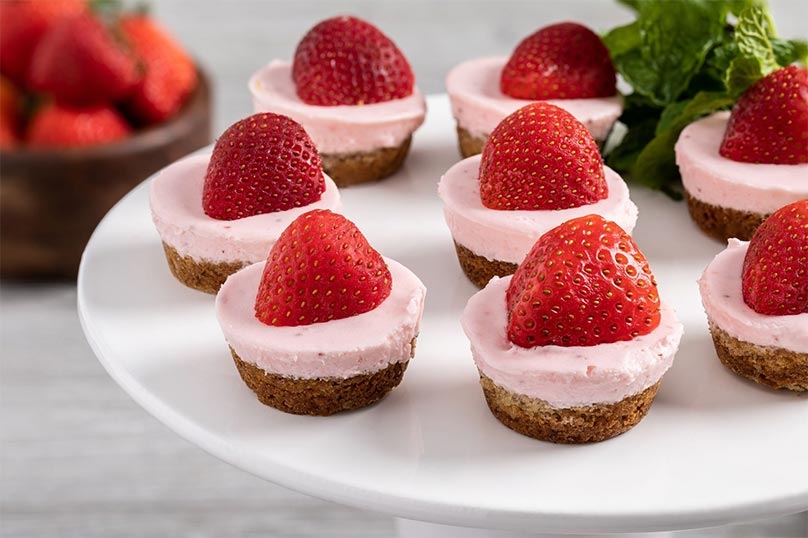 White serving plate with strawberry cheesecake cookie cups and mint leaves, stack of dessert plates, wooden bowl of strawberries, fresh mint leaves, gray wood table