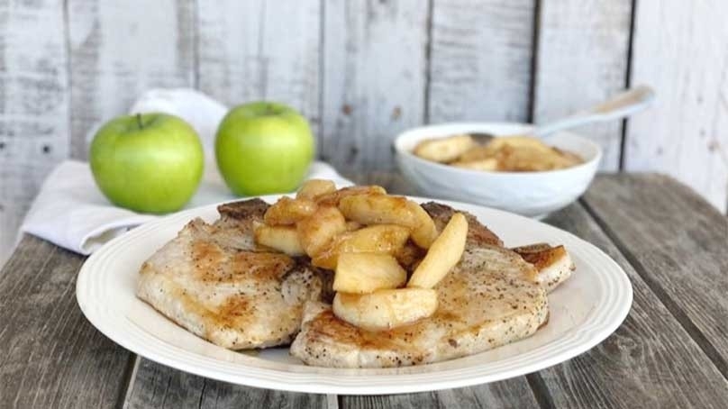 Skillet cinnamon apple pork chop on white plate topped with cooked apples, fresh green apples in background, wood table