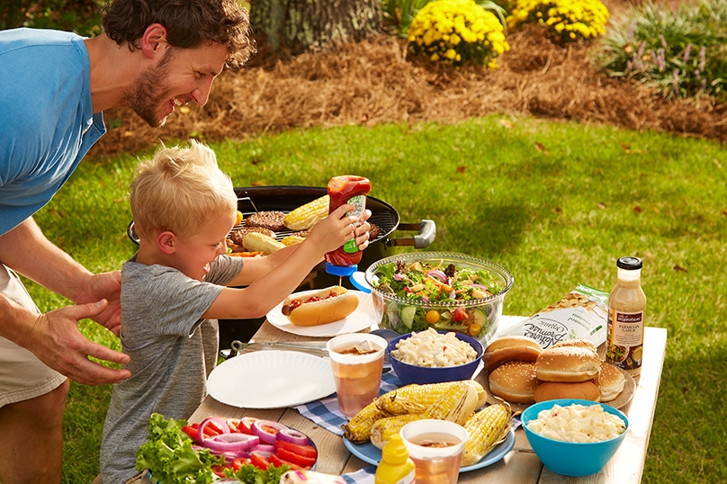 Father and son making hotdog at backyard barbecue, picnic table with food layout, grill with corn and burgers