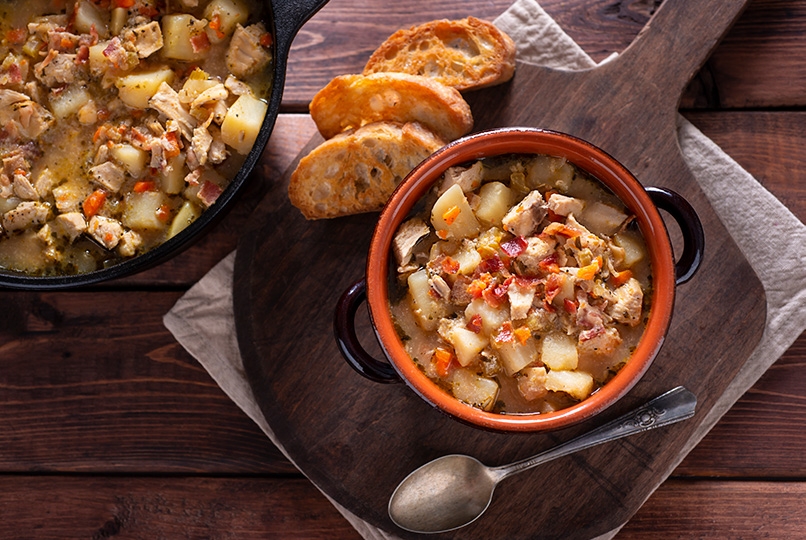 Top down view of bowl of hearty potato chicken soup with bacon on cutting board with toasted bread, spoon, gray napkin, cast iron pan of chicken stew in background