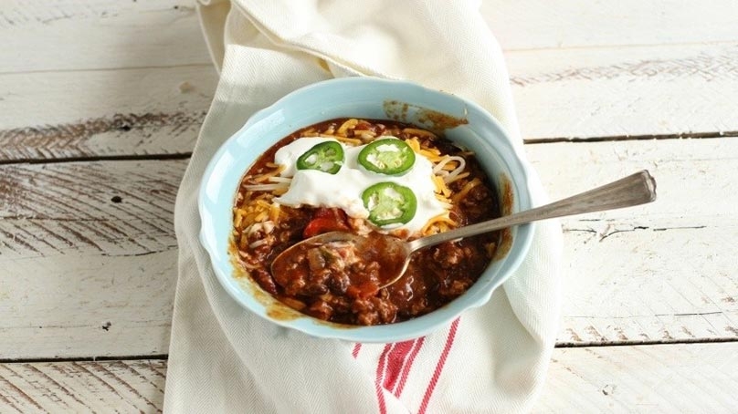 Spoon in Bowl of Chili topped with sour cream, cheese, and jalapeno slices, dinner napkin, white wood table