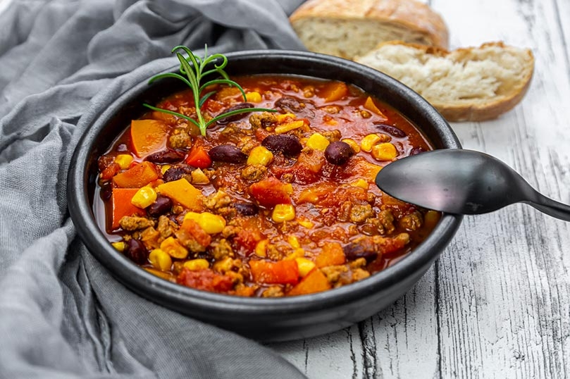 Mexican-style chili in black bowl, spoon, gray dinner napkin, bread roll, white-gray wood table