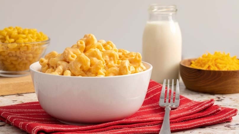  Slow Cooker Creamy Mac and Cheese  in white dish on red napkin with fork, cutting board, noodles, milk, shredded scheese