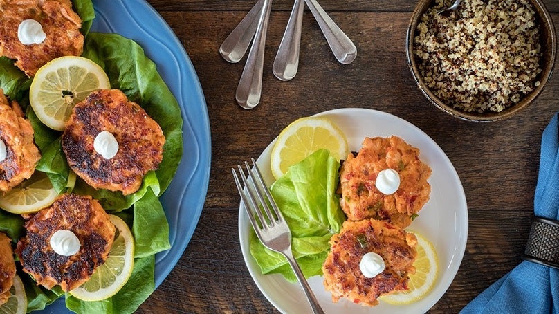 Spicy Salmon Cornmeal Cakes, blue serving platter, white plate, lemon slices forks, bowl of seasoning, blue napkin, dark wood table