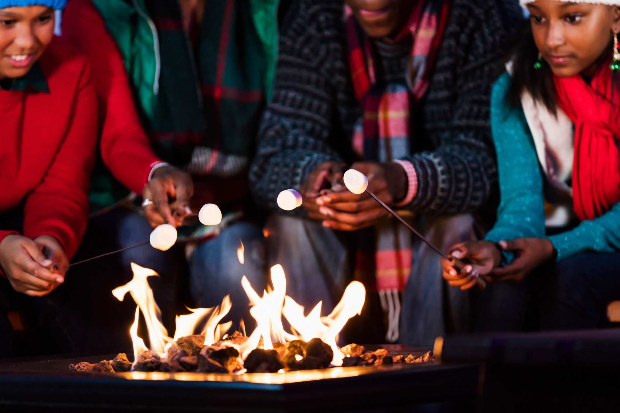 Family with two children gathered around fire pit, roasting marshmallows, wearing winter scarves and sweaters