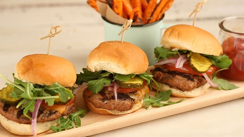 Three turkey burger sliders with pickles, red onion, tomato, parsley, serving dish of ketchup, serving bowl of sweet potato fries