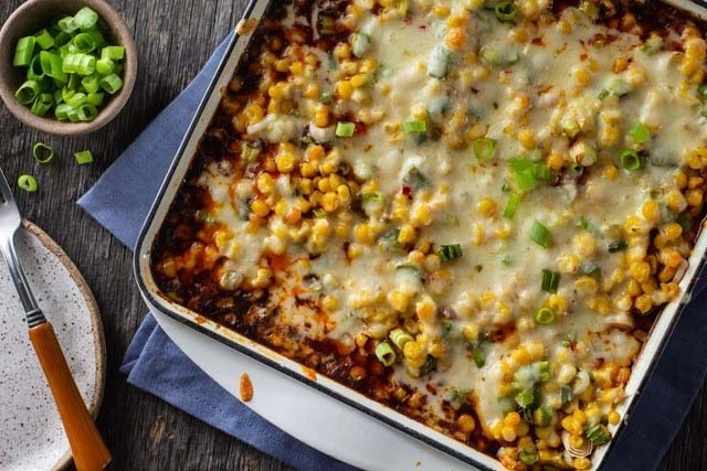 Chili Corn Casserole in baking pan, kitchen towel, plate, fork, cut gren onions in small bowl, rustic wood table